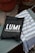 A black bag with white lettering displaying the brand name 'LUMI' and the phrase 'RELAX & RESTORE' is placed on a wooden bench. Next to the bag is a blue and white striped fabric, possibly a towel. The background features a concrete surface with a terracotta pot.