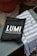 A black bag with white lettering displaying the brand name 'LUMI' and the phrase 'RELAX & RESTORE' is placed on a wooden bench. Next to the bag is a blue and white striped fabric, possibly a towel. The background features a concrete surface with a terracotta pot.