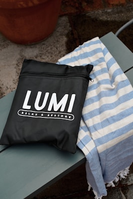 A black bag with white lettering displaying the brand name 'LUMI' and the phrase 'RELAX & RESTORE' is placed on a wooden bench. Next to the bag is a blue and white striped fabric, possibly a towel. The background features a concrete surface with a terracotta pot.