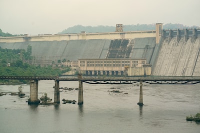 A large concrete dam features prominently, with a structure in the center possibly housing control equipment. The dam spans across a wide river, with water visible in the foreground. A bridge crosses the river, supported by multiple pillars. The surrounding landscape is lush with greenery and forested hillsides.