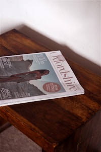 A magazine titled 'Oxfordshire Living' lies on a wooden table. The cover features an image of a person in an elegant outfit against a scenic outdoor backdrop. Headlines highlight topics such as quiet luxury, river cottage, and a festival.