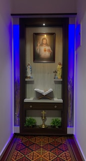 A small prayer altar features a portrait of Jesus with a sacred heart at the center. Below the portrait, two religious statues are positioned on either side, and a cross placed in the middle. An open book rests on a shelf below these items. The area is illuminated with soft blue lighting, and there are decorative elements such as plants and a patterned carpet.