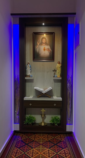 A small prayer altar features a portrait of Jesus with a sacred heart at the center. Below the portrait, two religious statues are positioned on either side, and a cross placed in the middle. An open book rests on a shelf below these items. The area is illuminated with soft blue lighting, and there are decorative elements such as plants and a patterned carpet.