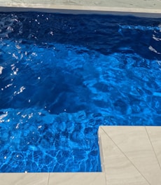 Rippling blue water in a swimming pool next to a light-colored stone or tile pool edge under bright sunlight.