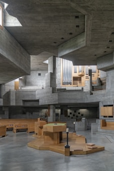 A modern, angular interior space made primarily of concrete with geometric architectural structures. Wooden elements add warmth to the overall gray tones, including benches and a central focal point resembling an altar. Large organ pipes are visible in the background, suggesting this is a church or a place of worship. The space has ample lighting with windows placed in high positions.