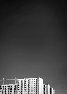 A black and white image features tall modern apartment buildings against a vast, dark sky. The buildings have a distinctive grid-like pattern of windows. Two cranes rise on either side, indicating ongoing construction. The overall composition directs attention to the architecture with the open sky enhancing the sense of height.