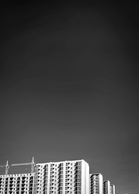 A black and white image features tall modern apartment buildings against a vast, dark sky. The buildings have a distinctive grid-like pattern of windows. Two cranes rise on either side, indicating ongoing construction. The overall composition directs attention to the architecture with the open sky enhancing the sense of height.