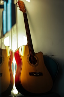 An acoustic guitar is leaning against a wall, beautifully illuminated by a warm, soft light. The wooden texture and the details of the guitar are clearly visible, and part of the guitar is reflected in a nearby mirror.