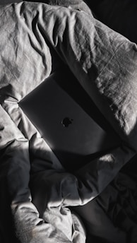 A dark-colored laptop with a notable logo is partially concealed by crumpled, textured bedsheets that have a gray hue. Sunlight casts dramatic shadows and highlights across the fabric and the laptop, creating a play of light and dark.