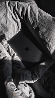 A dark-colored laptop with a notable logo is partially concealed by crumpled, textured bedsheets that have a gray hue. Sunlight casts dramatic shadows and highlights across the fabric and the laptop, creating a play of light and dark.