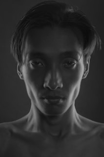 A black and white portrait of a person with short hair, looking directly at the camera. The lighting creates deep shadows and highlights the contours of the face, emphasizing the cheekbones and jawline. The expression is neutral and composed.