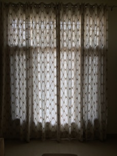 A set of floor-length curtains with a geometric pattern hangs in front of a window. The curtains are made of a semi-transparent fabric allowing diffused light to pass through, creating shadows on the floor.