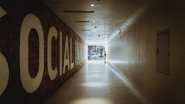 A long, tiled hallway with a large sign on the left reading 'SOCIAL INNOVATION,' leading towards a bright, glass doorway at the end. A person is standing near the doorway, interacting with something on the wall.