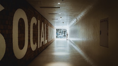 A long, tiled hallway with a large sign on the left reading 'SOCIAL INNOVATION,' leading towards a bright, glass doorway at the end. A person is standing near the doorway, interacting with something on the wall.