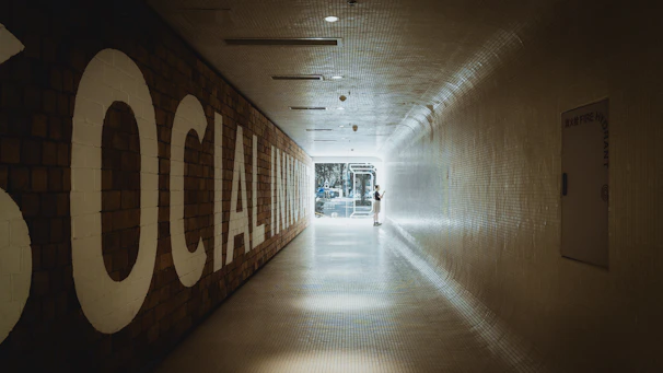 A long, tiled hallway with a large sign on the left reading 'SOCIAL INNOVATION,' leading towards a bright, glass doorway at the end. A person is standing near the doorway, interacting with something on the wall.