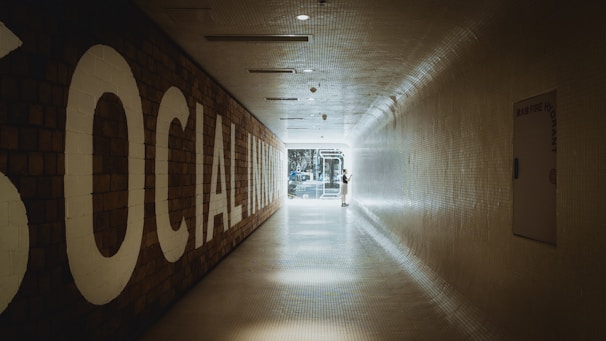 A long, tiled hallway with a large sign on the left reading 'SOCIAL INNOVATION,' leading towards a bright, glass doorway at the end. A person is standing near the doorway, interacting with something on the wall.