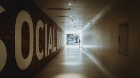 A long, tiled hallway with a large sign on the left reading 'SOCIAL INNOVATION,' leading towards a bright, glass doorway at the end. A person is standing near the doorway, interacting with something on the wall.