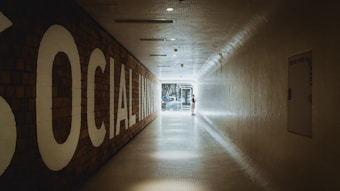 A long, tiled hallway with a large sign on the left reading 'SOCIAL INNOVATION,' leading towards a bright, glass doorway at the end. A person is standing near the doorway, interacting with something on the wall.