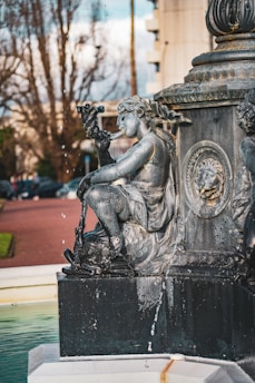 A detailed carving of a mythological figure is integrated into a decorative fountain. The figure appears to be holding a conch shell and is intricately designed with flowing garments and aquatic features, such as fish scales on the tail. Water is gently trickling from the sculpture, adding a dynamic element to the scene. In the background, there are blurred trees and urban elements, suggesting a park or plaza setting.