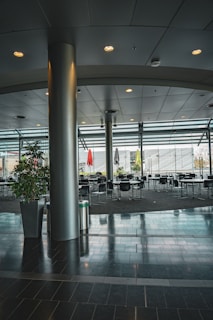 A spacious, modern interior with high ceilings and large windows allowing natural light to illuminate the space. The area is filled with several round tables and chairs, suggesting a cafeteria or dining area. Potted plants and colorful umbrellas add a touch of greenery and vibrancy.