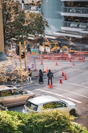 A city street scene with road construction, featuring multiple construction vehicles and barriers. Orange traffic cones and signs indicate an active work zone. Pedestrians are waiting at a crosswalk, and several parked cars are visible. There is a multi-story parking garage in the background and trees add greenery to the urban environment.