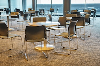 A modern, empty cafeteria area with round tables and sleek, black chairs arranged in an orderly fashion on a carpeted floor. Large windows provide ample natural light, highlighting the clean and minimalist design of the space.