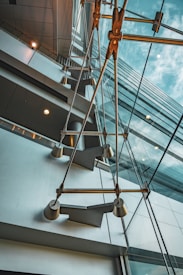 An abstract architectural view of a modern building interior featuring large glass windows and visible metal structures. The perspective gives a dramatic and dynamic upward angle, capturing reflections and intersecting lines between the glass panes and metallic elements.