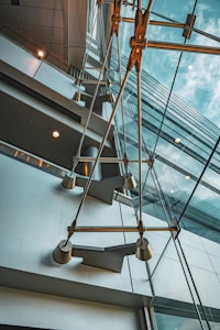 An abstract architectural view of a modern building interior featuring large glass windows and visible metal structures. The perspective gives a dramatic and dynamic upward angle, capturing reflections and intersecting lines between the glass panes and metallic elements.