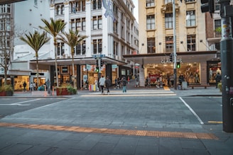 A busy urban street scene with several people crossing at a crosswalk. Pedestrian traffic lights are visible with a green signal. Multiple storefronts, including shops with sale signs and clothing displays, line the modern buildings. Tall palm trees are planted along the sidewalk, adding a touch of greenery. The setting suggests a bustling city environment with commercial activity.