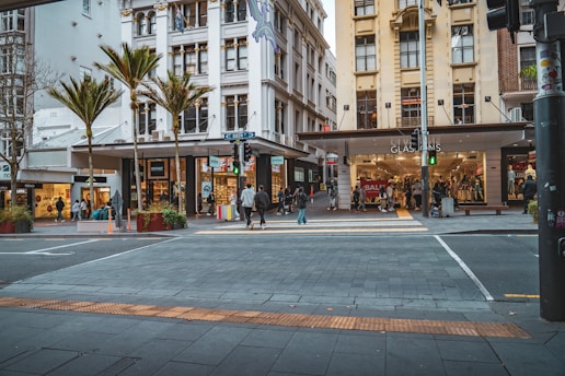 A busy urban street scene with several people crossing at a crosswalk. Pedestrian traffic lights are visible with a green signal. Multiple storefronts, including shops with sale signs and clothing displays, line the modern buildings. Tall palm trees are planted along the sidewalk, adding a touch of greenery. The setting suggests a bustling city environment with commercial activity.