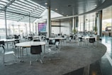 This modern, spacious indoor area features multiple round tables with black chairs arranged around them. The floor is carpeted, and floor-to-ceiling windows allow ample natural light to illuminate the space. There are potted plants around the room, and a large digital screen is visible in the background. The ceiling features recessed lighting and metal pillars.