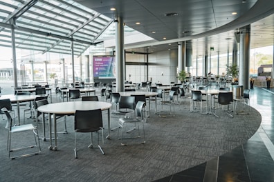 This modern, spacious indoor area features multiple round tables with black chairs arranged around them. The floor is carpeted, and floor-to-ceiling windows allow ample natural light to illuminate the space. There are potted plants around the room, and a large digital screen is visible in the background. The ceiling features recessed lighting and metal pillars.