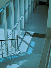 A modern staircase with metallic railings is bathed in a blue-green light, creating geometric shadows on the tiled floor. The scene is framed by large, vertical glass windows, providing a sleek and minimalist architectural impression.