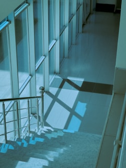 A modern staircase with metallic railings is bathed in a blue-green light, creating geometric shadows on the tiled floor. The scene is framed by large, vertical glass windows, providing a sleek and minimalist architectural impression.