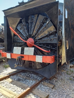 A close-up view of a snowplow train attachment, featuring large rotating blades and a central red hub. Two informational plaques are positioned near the bottom, and the apparatus is mounted on railroad tracks. The machinery has a weathered appearance, with visible metal parts and some rust.