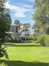 A large, well-maintained house with white windows is surrounded by lush greenery and trees. The expansive lawn is bright green, and a smaller building is visible in the background. The sky is blue with scattered white clouds, creating a serene and peaceful setting.