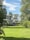 A large, well-maintained house with white windows is surrounded by lush greenery and trees. The expansive lawn is bright green, and a smaller building is visible in the background. The sky is blue with scattered white clouds, creating a serene and peaceful setting.