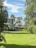 A large, well-maintained house with white windows is surrounded by lush greenery and trees. The expansive lawn is bright green, and a smaller building is visible in the background. The sky is blue with scattered white clouds, creating a serene and peaceful setting.