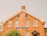 A brick building with white-framed windows and a gabled roof, housing the Amana Heritage Museum and Book Store. The sign over the door indicates the museum entrance. The building is partially obscured by greenery in the foreground.