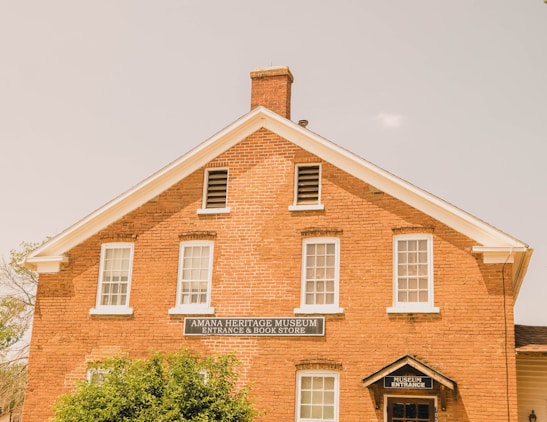 A brick building with white-framed windows and a gabled roof, housing the Amana Heritage Museum and Book Store. The sign over the door indicates the museum entrance. The building is partially obscured by greenery in the foreground.
