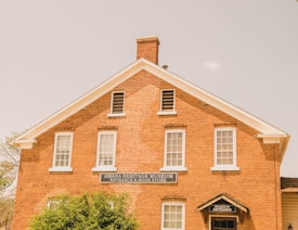 A brick building with white-framed windows and a gabled roof, housing the Amana Heritage Museum and Book Store. The sign over the door indicates the museum entrance. The building is partially obscured by greenery in the foreground.