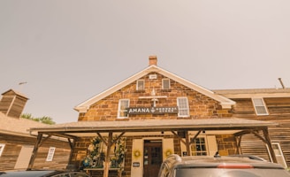 A rustic general store with a brick facade, featuring a gabled roof with a vintage sign that reads 'Amana General Store'. The front area has wooden support beams and is decorated with planters holding flowers and wreaths. Several cars are parked in front, and the sunlight creates a warm, sepia-toned atmosphere.