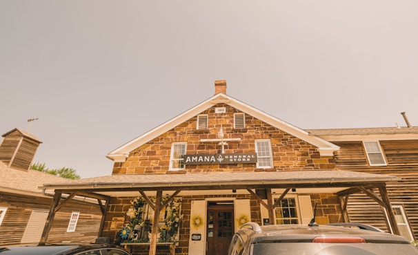 A rustic general store with a brick facade, featuring a gabled roof with a vintage sign that reads 'Amana General Store'. The front area has wooden support beams and is decorated with planters holding flowers and wreaths. Several cars are parked in front, and the sunlight creates a warm, sepia-toned atmosphere.