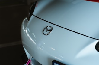 The image features the front part of a white car with a prominent Mazda logo on the hood. The setting seems to be a dimly lit garage or parking space, with subtle reflections on the vehicle's surface indicating overhead lighting.
