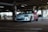 A white sports car with custom graphics is parked in a dimly lit parking garage. The car features a colorful design on its side, incorporating shades of pink and purple. It's equipped with dark alloy wheels and a front bumper tow hook.