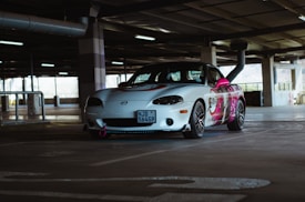 A white sports car with custom graphics is parked in a dimly lit parking garage. The car features a colorful design on its side, incorporating shades of pink and purple. It's equipped with dark alloy wheels and a front bumper tow hook.