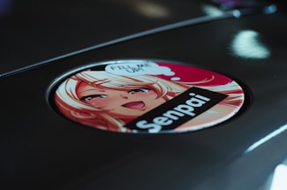 A circular sticker featuring an anime-style illustration of a blonde character with bright eyes and a smiling expression. The character is accompanied by speech bubbles and text, with a bold label reading 'Senpai.' The sticker is placed on a reflective metallic surface.