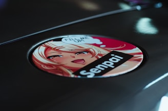 A circular sticker featuring an anime-style illustration of a blonde character with bright eyes and a smiling expression. The character is accompanied by speech bubbles and text, with a bold label reading 'Senpai.' The sticker is placed on a reflective metallic surface.
