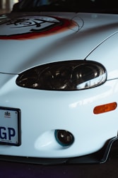 Close-up of the front of a white car with a prominent headlight, a bumper with an orange indicator light, a license plate reading 'GP', and a decal with a stylized design in red and black.