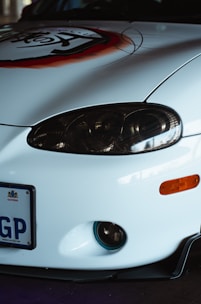 Close-up of the front of a white car with a prominent headlight, a bumper with an orange indicator light, a license plate reading 'GP', and a decal with a stylized design in red and black.